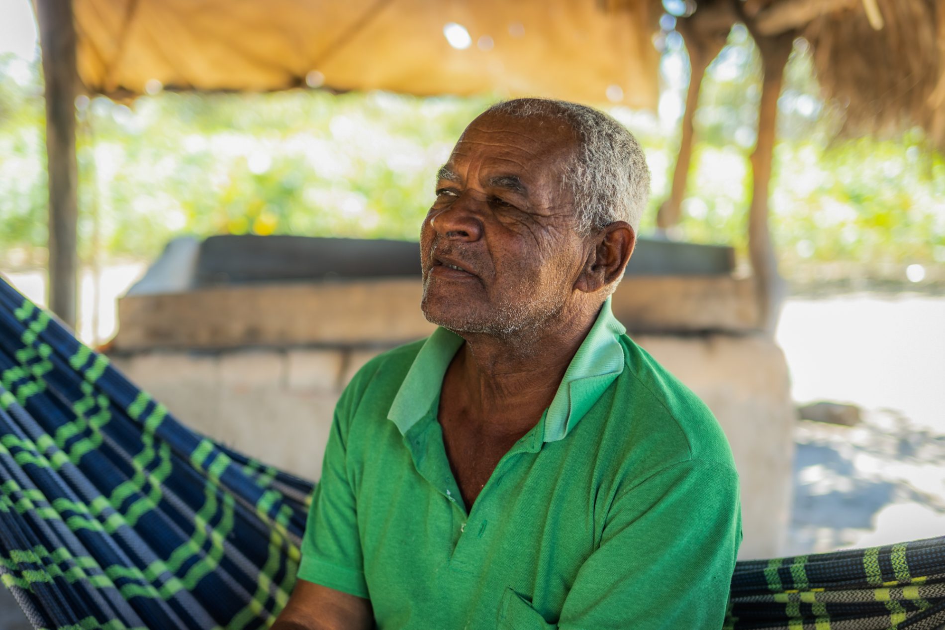 Retrato de um senhor negro de cabelos brancos sentado em uma rede com uma camisa gola polo verde sob uma construção simples, sem paredes, com teto de palha.
