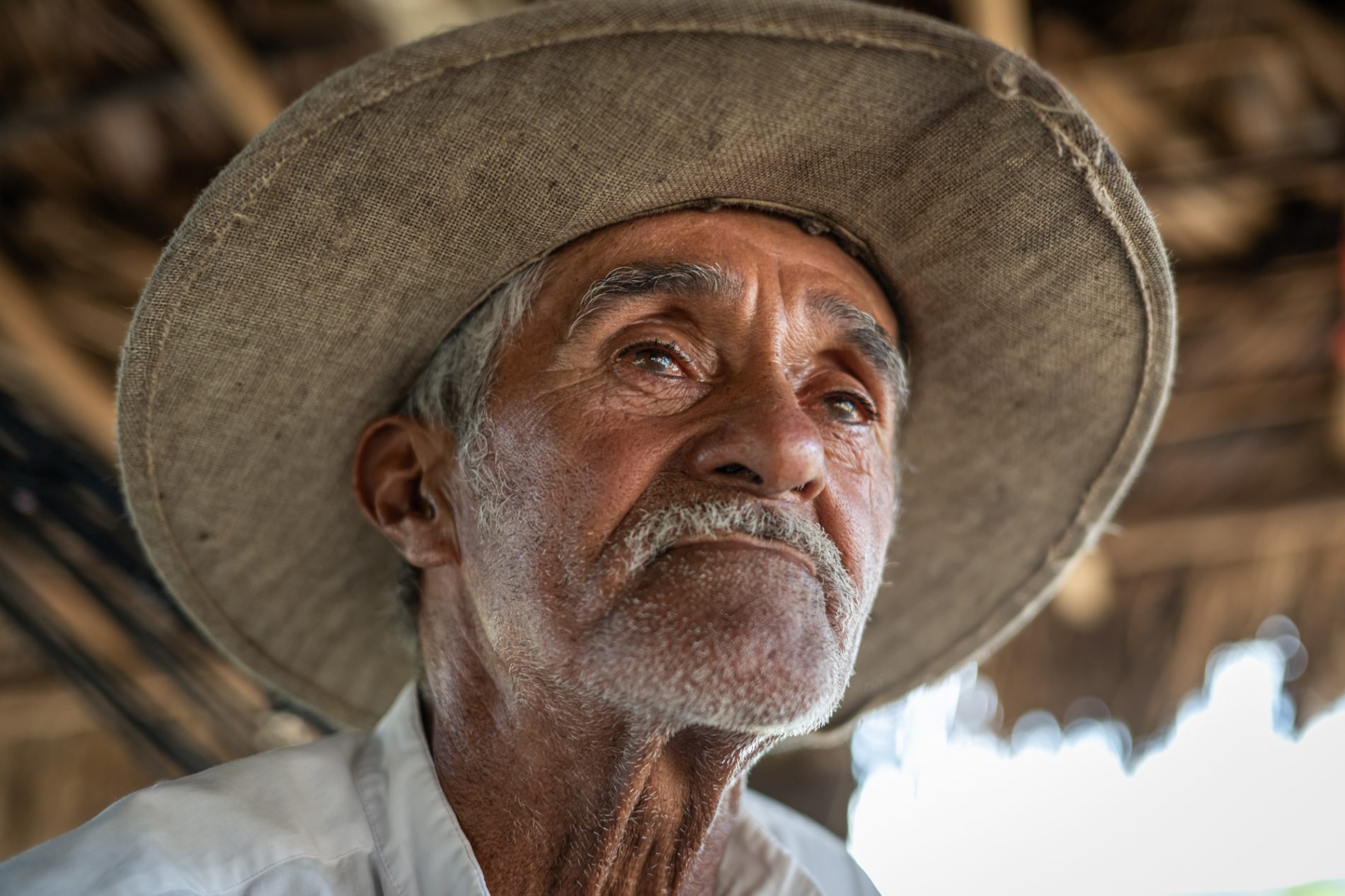 Retrato de senhor de cabelos e bigode brancos, com um chapéu e sob uma construção simples, sem paredes, com teto de palha.