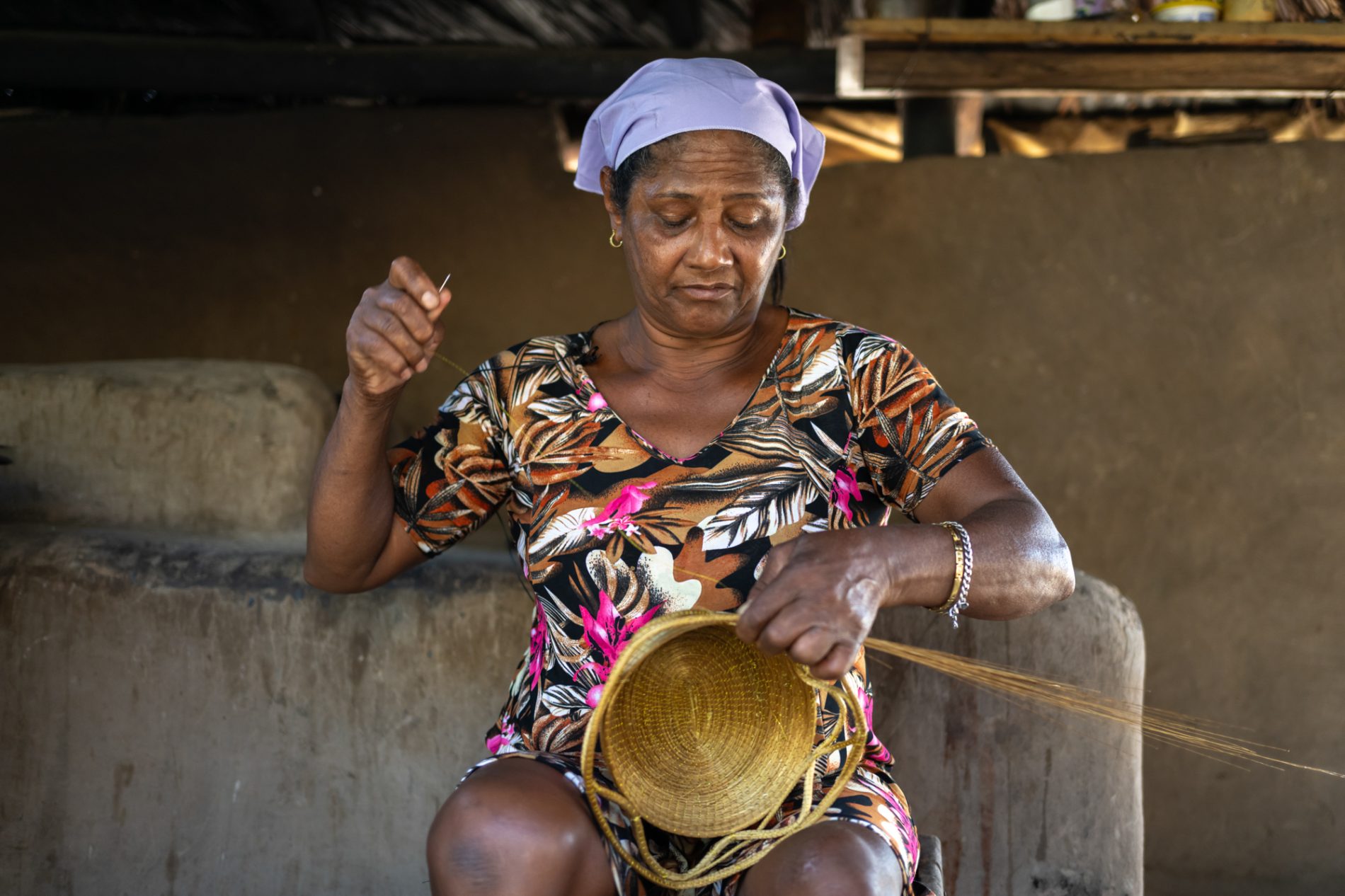 Retrato de uma senhora segurando uma agulha em uma mão e um cesto em outra, dentro de uma construção simples, com teto sem forro e paredes sem pintura.