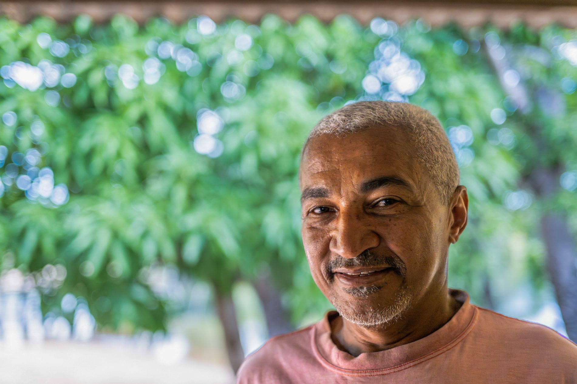 Retrato de senhor de cabelos e bigode brancos, com um chapéu e sob uma construção simples, sem paredes, com teto de palha.