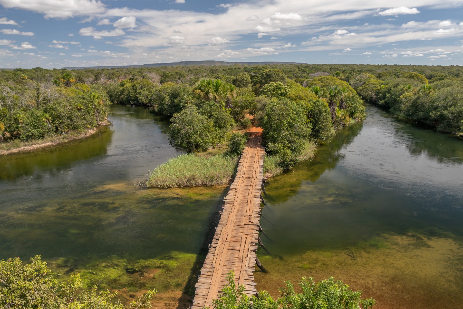 Foto aérea mostra uma ponte de madeira sobre um rio de águas transparentes esverdeadas. Ao redor, vegetação preservada de Cerrado.