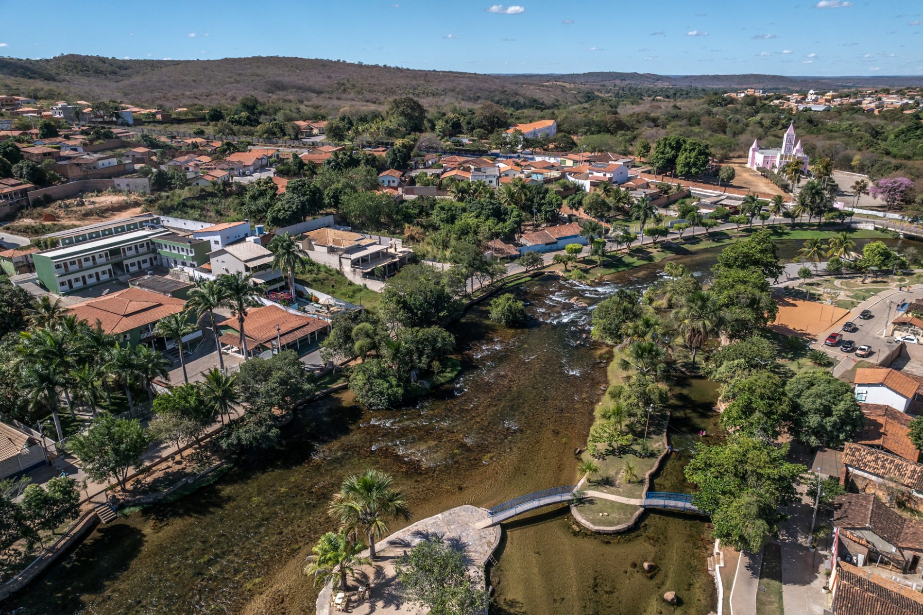 Vista aérea de uma cidade pequena com casas de telhados vermelhos, áreas verdes e ruas tranquilas. Um rio de águas rasas e claras atravessa o centro urbano, com pequenas quedas d’água, ilhotas e vegetação ciliar. Há passarelas e calçadas margeando o rio, além de palmeiras e árvores oferecendo sombra. Ao fundo, surgem morros cobertos por vegetação seca típica do Cerrado. No lado direito, destaca-se uma igreja grande de fachada rosa claro.