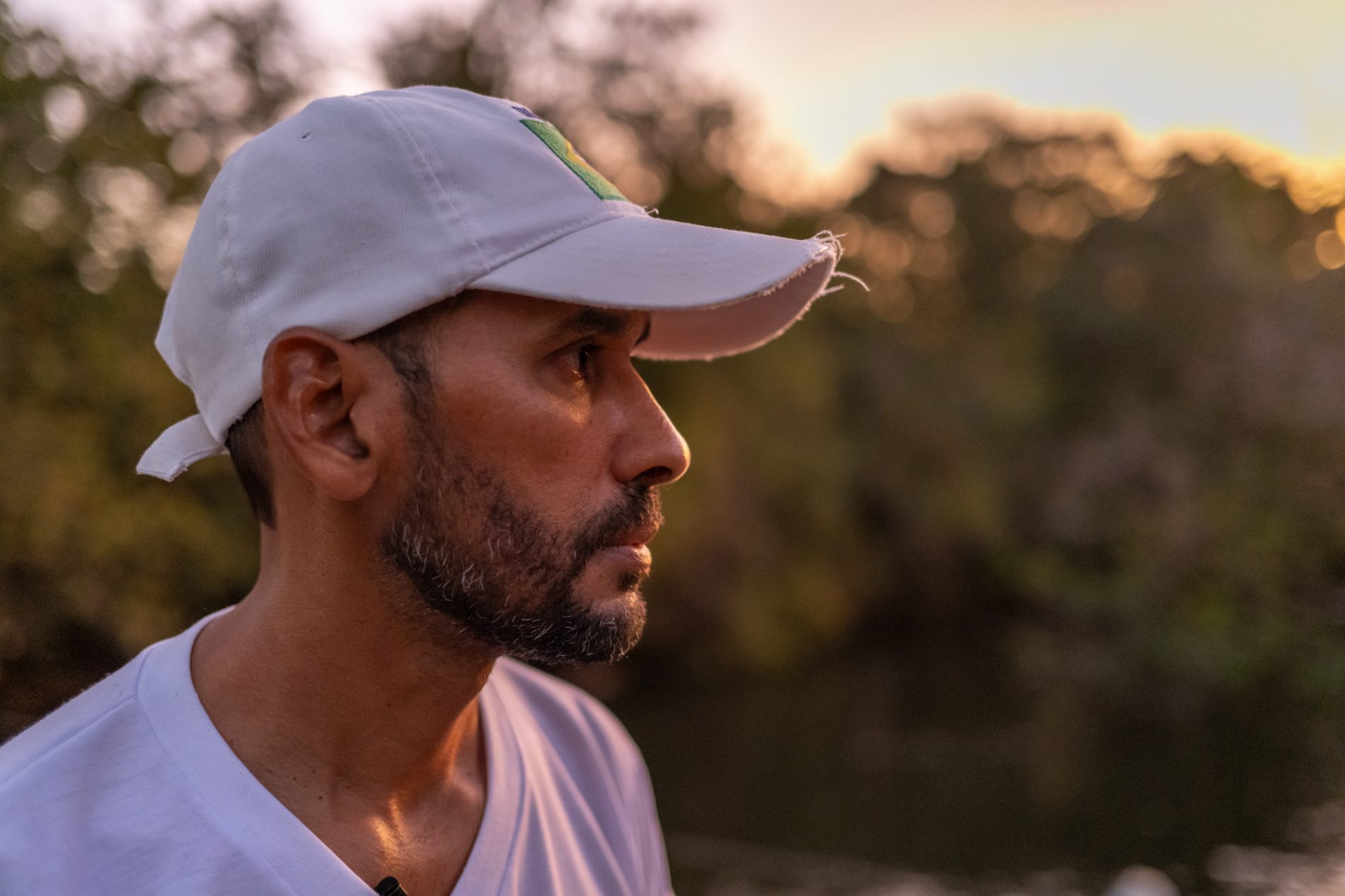 Homem de meia-idade com boné branco e camiseta clara, visto em perfil ao entardecer. Ao fundo, a vegetação está desfocada, com luz suave do pôr do sol. Ele parece observador e reflexivo.