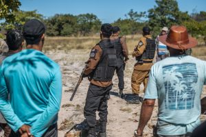 Grupo de policiais armados com coletes à prova de balas confronta homens vestidos com roupas civis em uma área rural. A cena mostra tensão entre os dois grupos, que estão em um espaço aberto de vegetação rasteira e árvores ao fundo.