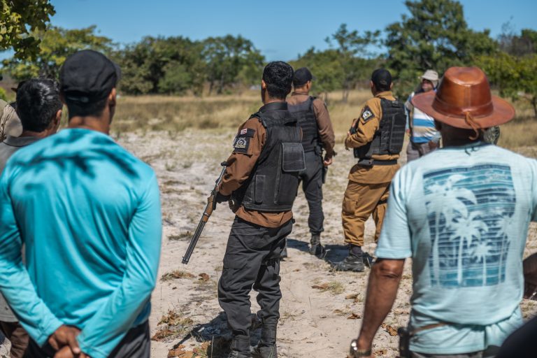 Grupo de policiais armados com coletes à prova de balas confronta homens vestidos com roupas civis em uma área rural. A cena mostra tensão entre os dois grupos, que estão em um espaço aberto de vegetação rasteira e árvores ao fundo.