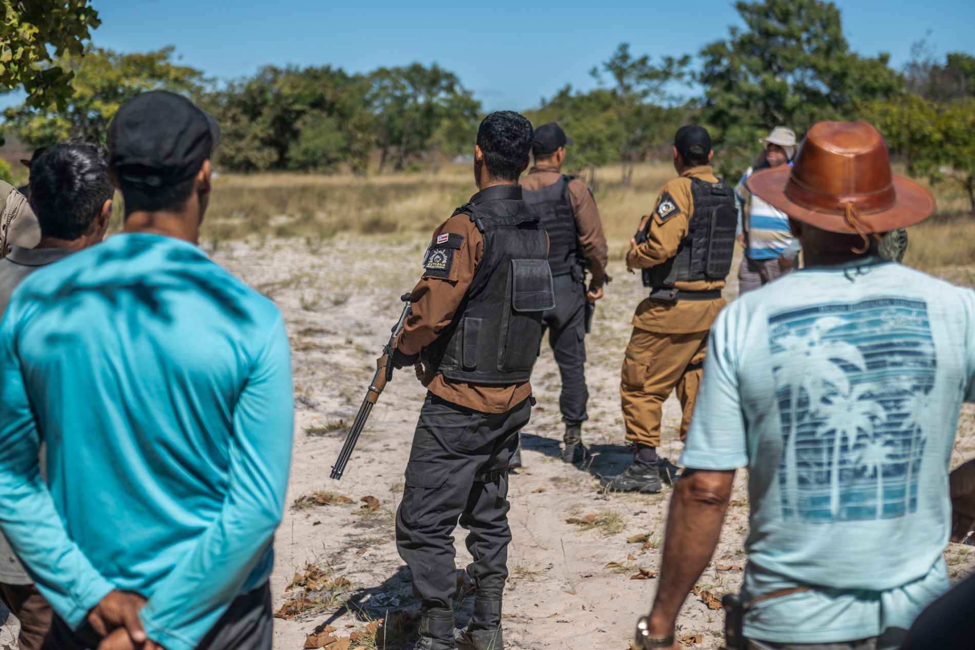 Grupo de policiais armados com coletes à prova de balas confronta homens vestidos com roupas civis em uma área rural. A cena mostra tensão entre os dois grupos, que estão em um espaço aberto de vegetação rasteira e árvores ao fundo.