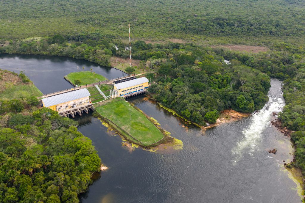 Imagem aérea de usina hidrelétrica, que aparece no lado esquerdo, região central da imagem, na cor branca; ao redor, rio e vegetação.