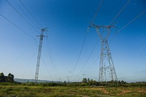 Torres de alta tensão em área rural, com cabos elétricos atravessando o céu azul sobre vegetação baixa e colinas ao fundo.