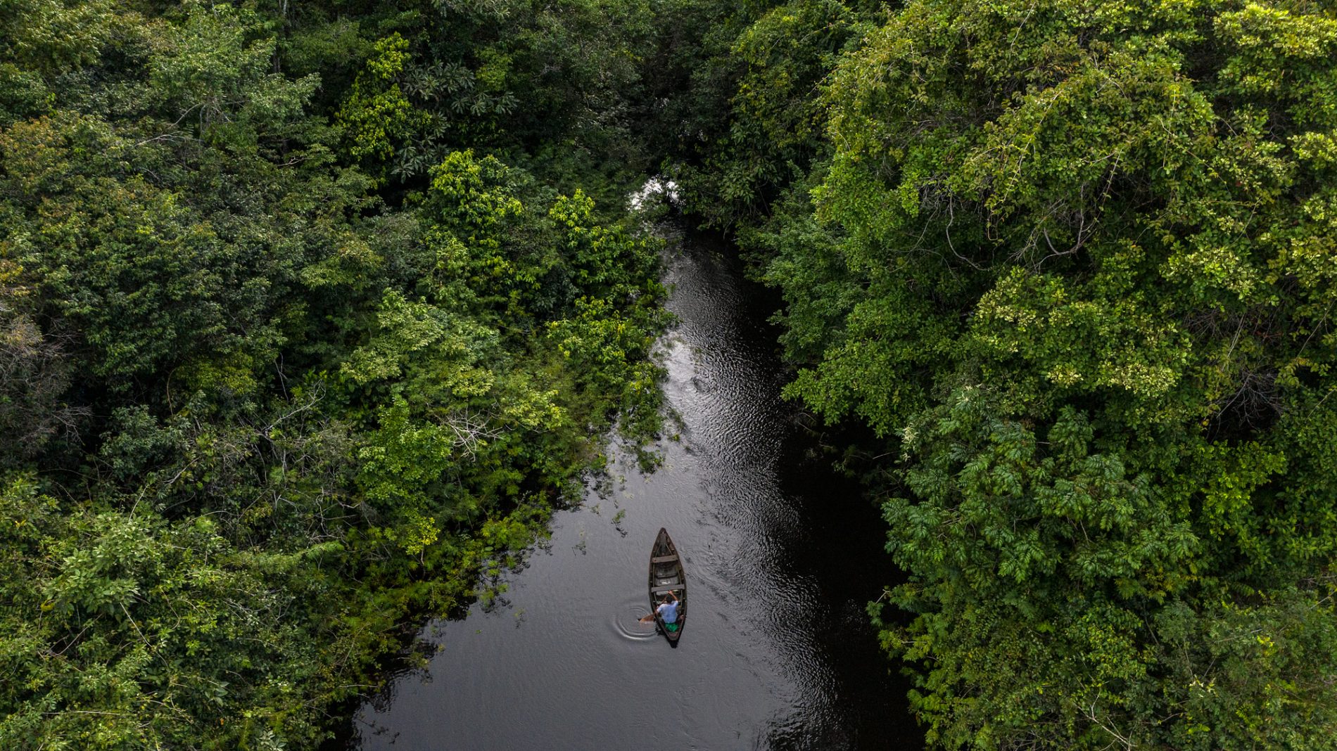 Vista aérea de um igarapé estreito cercado por floresta densa, com uma pequena canoa e duas pessoas navegando pelo centro do curso d’água.