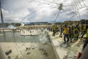 Vidro estilhaçado em primeiro plano, com marcas de impactos e um grande buraco, revela ao fundo uma multidão de manifestantes vestidos de verde e amarelo, alguns com bandeiras do Brasil, durante os ataques golpistas de 8 de janeiro de 2023, na Praça dos Três Poderes, em Brasília.