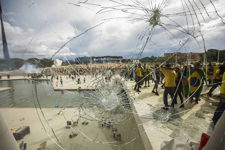 Vidro estilhaçado em primeiro plano, com marcas de impactos e um grande buraco, revela ao fundo uma multidão de manifestantes vestidos de verde e amarelo, alguns com bandeiras do Brasil, durante os ataques golpistas de 8 de janeiro de 2023, na Praça dos Três Poderes, em Brasília.